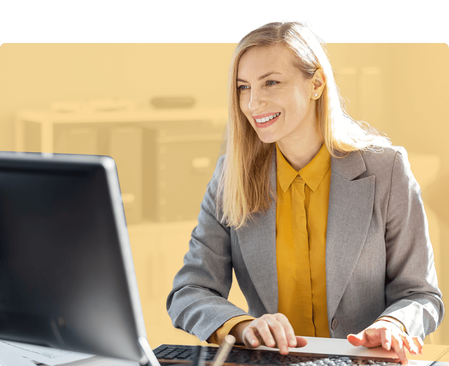 Eine Frau mit langen blonden Haaren und einem Lächeln im Gesicht bedient eine Tastatur an einem Arbeitsplatz. Sie trägt ein graues Sakko und eine gelbe Bluse. Im Hintergrund ist ein unscharfer Bürobereich zu erkennen.