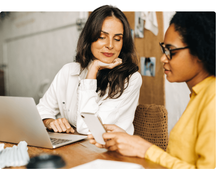 Zwei Frauen bei einer Besprechung, eine nutzt einen Laptop, die andere ein Smartphone.