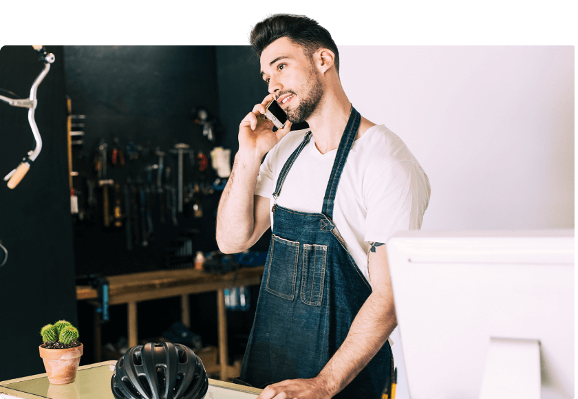 Ein junger Mann in einer Werkstatt trägt eine Schürze und spricht am Telefon. Er steht vor einem Computer, während im Hintergrund Werkzeuge und Fahrradzubehör zu sehen sind, was auf ein handwerkliches Geschäft hinweist.