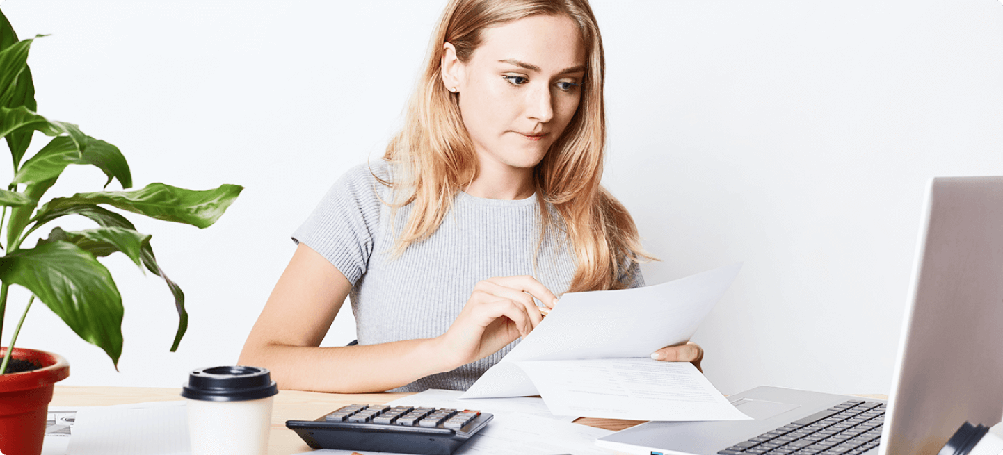 Junge Frau in einem grauen T-Shirt, die konzentriert an einem Schreibtisch sitzt, einen Papierbeleg in der Hand hält und mit einem Laptop arbeitet, während ein Taschenrechner und eine Tasse Kaffee neben ihr liegen.