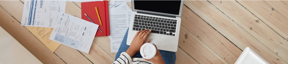 Nahaufnahme einer Person, die mit einem Laptop arbeitet und dabei eine Tasse Kaffee hält, umgeben von Rechnungen und Dokumenten auf einem Holzboden.