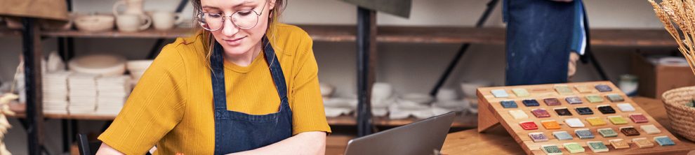 Junge Frau mit Brille und gelbem Shirt, die in einer Werkstatt am Laptop arbeitet und Notizen macht, umgeben von Arbeitsmaterialien.